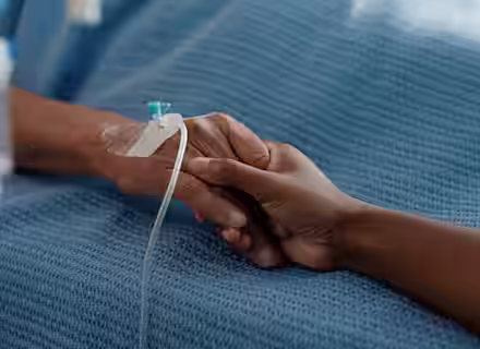 A close-up of two people holding hands; one person is lying in a hospital bed with an IV in their hand, while the other offers comfort and support.