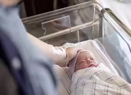 A newborn baby wrapped in a blanket sleeps in a hospital bassinet while an adult hand gently rests on the baby's head. The adult has a hospital ID band on their wrist.