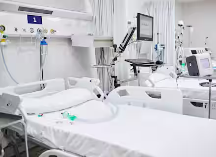A hospital room with multiple empty, neatly made hospital beds. Medical equipment such as monitors, ventilators, and IV stands are beside the beds. Everything is clean and organized, creating a sterile environment. Curtains provide privacy between beds.