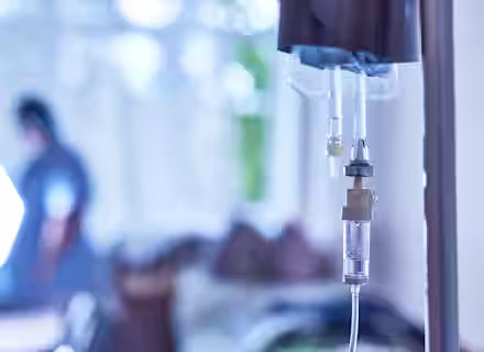 Close-up of an intravenous drip chamber and tubing, with a blurred figure of a person in scrubs in the background. The setting appears to be a hospital room with bright, natural light coming through a window.