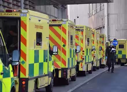 A row of NHS ambulances with yellow and green markings are parked outside a hospital. A paramedic in uniform walks along the pavement beside the vehicles.