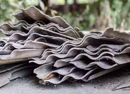 A close-up view of several stacked, weathered, and broken corrugated asbestos cement roofing sheets with a blurred outdoor background.