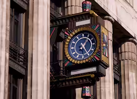A colorful, ornate clock with a blue face and gold hands is mounted on the exterior of a stone building with columns. The clock features geometric, multicolored patterns and stands out against the building's neutral facade.