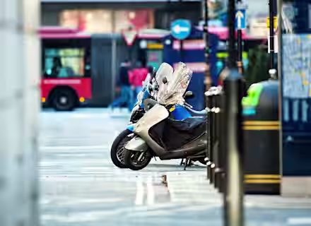Parked motorcycles on a city street with a red bus and pedestrians in the background. The scene includes street signs and a map on the right, with bollards lining the sidewalk.