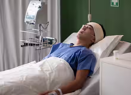 A man with a bandaged head lies in a hospital bed, wearing a blue gown and an oxygen tube. Medical equipment is nearby, and he looks up, appearing alert but serious.