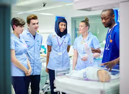 A group of four medical students in blue uniforms attentively listen to a healthcare professional explaining a procedure on an infant manikin in a hospital training room.