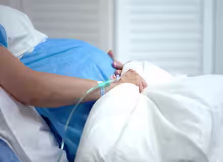 A pregnant person lying in a hospital bed, wearing a blue gown. Their hand is resting on their belly and has an IV line attached. The background consists of white curtains, and the scene suggests a medical or hospital setting.