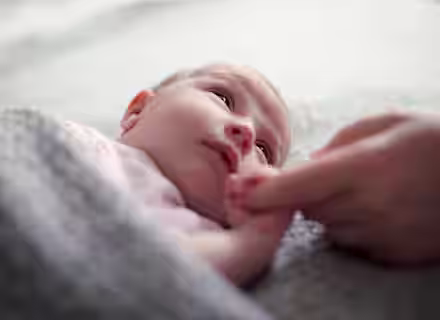 A baby lies on a soft, grey blanket, looking up. The baby is wrapped up in the blanket, and a person’s hand is gently holding the baby’s hand. The scene evokes a sense of warmth and tenderness.