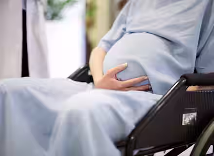 A pregnant person in a light blue hospital gown sits in a wheelchair, gently cradling their baby bump with one hand. A healthcare professional, partially visible and dressed in white, stands nearby. The background is softly blurred.