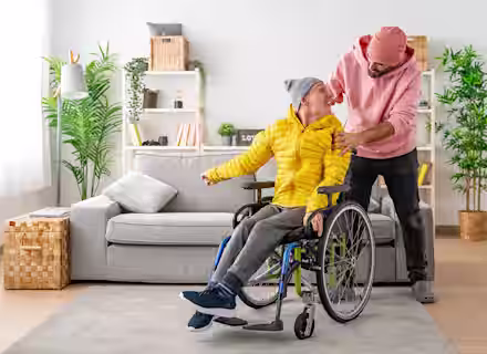 A smiling man in a yellow jacket sits in a wheelchair while another man in a pink hoodie and beanie stands behind him, both looking at each other happily in a bright living room.