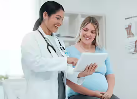 A doctor in a white coat shows information on a tablet to a smiling pregnant woman sitting beside her in a medical exam room.