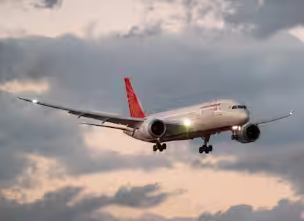 A commercial airplane with Air India livery is flying against a cloudy sky during sunset, with its landing gear extended and lights on.