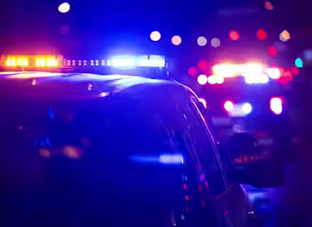 A night scene featuring several police vehicles with flashing blue and red lights illuminating the dark surroundings. The focus is on the lights of the police cars, creating a sense of urgency or alert. The background is blurred with additional flashing lights visible.