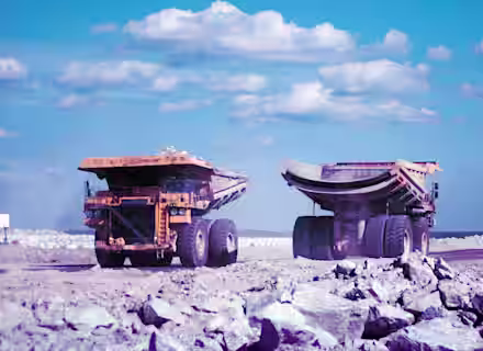 Two large heavy-duty trucks positioned on a rocky construction or mining site. The trucks, facing away from each other, show dusty tires and an open bed, surrounded by a landscape of rocks under a blue sky with scattered clouds.