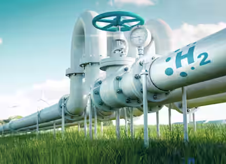 A metal pipeline marked with "H2" and a gauge is shown running through a green grassy field. Wind turbines are visible in the background under a clear blue sky, indicating renewable energy.