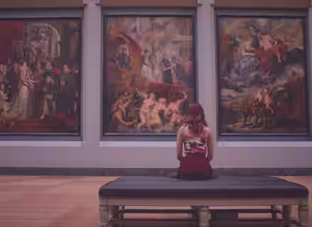 A person with shoulder-length hair, wearing a sleeveless top and skirt, sits on a bench in an art museum. They are facing three large classical paintings with intricate, vivid scenes, depicting numerous figures and actions in rich colors.