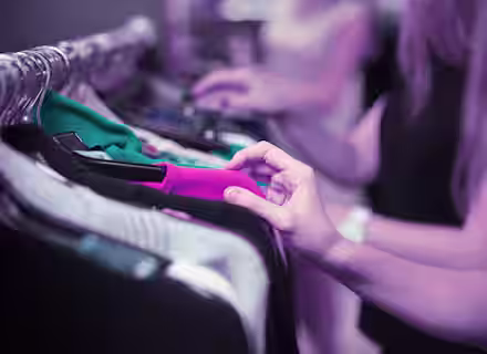 Close-up of people browsing through clothes on a rack. One hand is prominently seen touching a pink garment, while other blurred figures in the background also look through clothing items. The scene appears to be in a store or a market with a purple-tinted lighting.