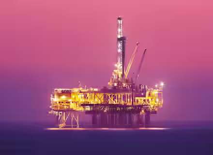 An offshore oil rig brightly lit against a serene dusk sky with a gradient from pink to orange. The rig stands alone in the middle of the calm ocean, with cranes and machinery visible. The water reflects the orange lights from the structure.