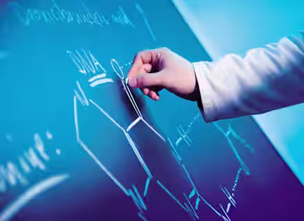 Close-up of a person's hand in a lab coat drawing a chemical structure on a chalkboard with chalk. The chalkboard has various scientific notations and diagrams related to DNA. The background is lit with a bluish hue.