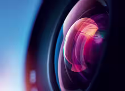 Close-up shot of a camera lens reflecting colorful light. The lens exhibits a spectrum of colors including pink, purple, and blue, with a blurred, gradient blue background that transitions from dark to light. The image emphasizes the intricate details of the lens.