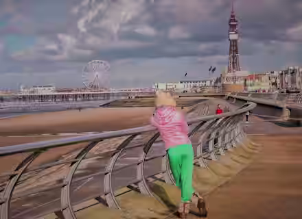 Person in a pink hoodie and green pants leans on a railing facing a beach. A Ferris wheel and tower are visible in the background against a cloudy sky. Other people are seen walking along the promenade.