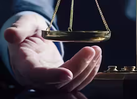 A person’s hand reaches toward a brass balance scale on a dark reflective surface, symbolizing justice and fairness, with a dark, blurred background.