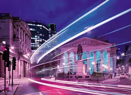 A nighttime city scene with light trails from passing vehicles, illuminating a historical building with columns and a statue in front. The sky is a deep blue, and the surrounding modern buildings are lit up, contrasting the neoclassical architecture.