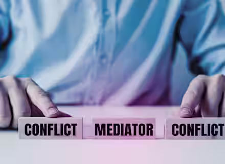 A person in a suit is seen from the torso down, pointing at three wooden blocks on a table. The blocks are labeled "CONFLICT," "MEDIATOR," and "CONFLICT." The image symbolizes the role of a mediator in resolving conflicts between two parties.
