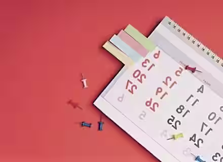 A desk calendar turned to the month of March, with several colored sticky notes attached to the top. Red, blue, yellow, and white thumbtacks are scattered on the red surface around the calendar.