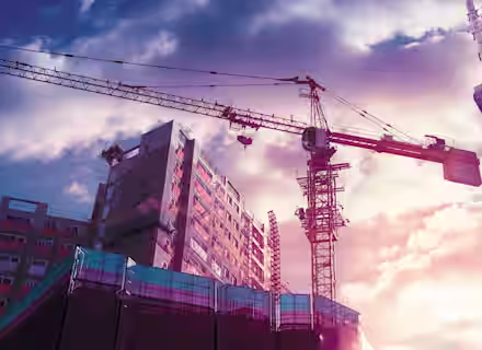 A construction site with tall cranes and buildings under construction. The sky is filled with dramatic clouds illuminated by a colorful sunset, casting a warm glow on the scene. Scaffolding and partially completed structures are prominent in the foreground.