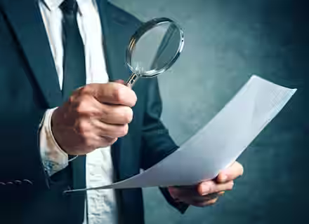 A person wearing a suit and tie is holding a magnifying glass in their right hand and examining a sheet of paper in their left hand. The background is blurred and has a dark, textured appearance.