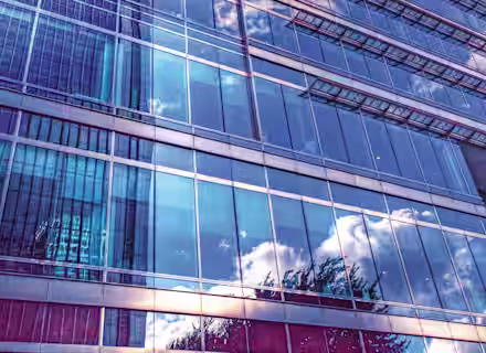 A modern skyscraper with a glass facade reflects the sky, clouds, and neighboring buildings. The image is taken from a low angle, emphasizing the height and sleek design of the structure. The blue tones create a serene, contemporary atmosphere.
