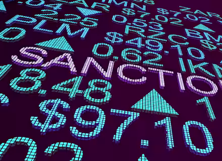 A digital display shows various currency symbols, numbers, and the words "SANCTIONS" prominently in the center. The background is a deep purple, with blue and green elements highlighting the text and symbols, suggesting a financial or economic theme.