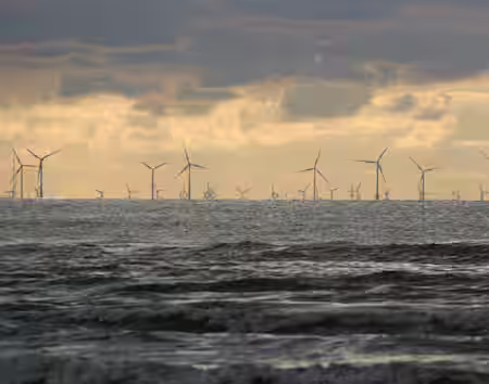 A broad view of numerous wind turbines on the horizon stretches across the ocean. The sky is cloudy with a backdrop of bright yellow and orange hues from the sunset, and the sea has gentle waves near the shore.