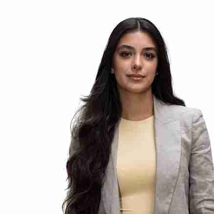 A woman with long, dark hair wearing a beige blazer and a light yellow top stands against a plain white background. She is looking directly at the camera with a neutral expression.