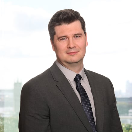 A man in a dark gray suit, light gray shirt, and dark polka dot tie is standing in front of a window with a blurred cityscape in the background. He has short dark hair and a slight smile, with hands clasped in front.