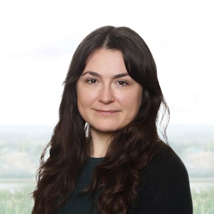 A woman with long, dark brown hair and fair skin is smiling slightly while looking directly at the camera. She is wearing a green top and a black cardigan. The background shows a blurred outdoor scene with cloudy skies and green fields.