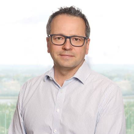 A man with short, dark hair and glasses is wearing a light blue, button-up striped shirt. He stands in front of a blurred outdoor background, looking directly at the camera with a neutral expression.