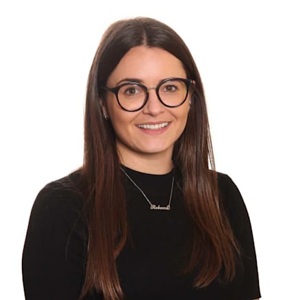 A woman with long straight brown hair, wearing black-rimmed glasses and a black top. She is smiling and wearing a necklace with a name pendant. The background is plain white.