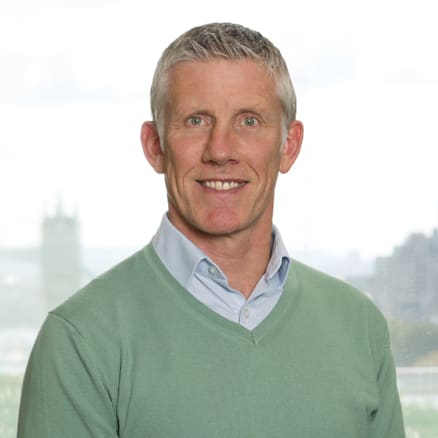 A man with short gray hair smiles for the camera. He is wearing a light green sweater over a light blue collared shirt. There is a cityscape with a tall building visible in the blurred background.