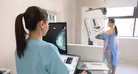 A healthcare professional in scrubs is viewing a digital mammogram on a screen while a patient in a hospital gown undergoes a mammogram examination in the background. The room is well-lit with medical equipment and a clipboard on a counter.