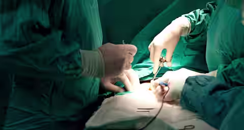 Two surgeons in green sterile gowns and gloves are performing surgery. They are focused on an incision, using surgical instruments to operate. The operating table is covered with a green drape, and additional tools are visible next to the patient.