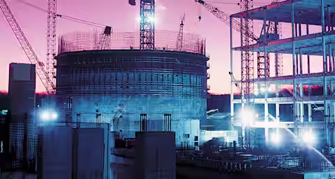 A construction site at dusk with illuminated cranes and structures. The sky is a gradient of pink and purple hues. Several cranes are actively working on a large cylindrical building in progress, surrounded by scaffolding and other partially built structures.