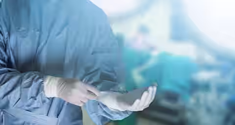 A surgeon wearing a blue surgical gown, mask, and gloves is preparing in the foreground, while an operating room with blurred figures and surgical equipment is visible in the background under bright lights.