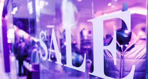 A store window with large white letters spelling "SALE" in a brightly lit retail environment. Inside, blurred mannequins display winter jackets. The scene has a purple-tinted lighting effect.
