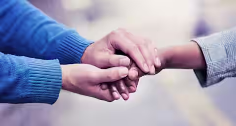 Two people holding hands tenderly. One person is wearing a blue sweater, and the other is wearing a gray jacket. The focus is on their hands, with a blurred background showing an outdoor setting. The gesture suggests comfort or support.
