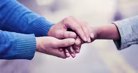 Two people holding hands tenderly. One person is wearing a blue sweater, and the other is wearing a gray jacket. The focus is on their hands, with a blurred background showing an outdoor setting. The gesture suggests comfort or support.