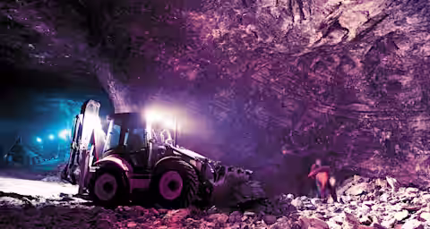 A large excavator operates within a dimly lit, rocky underground cave. The vehicle's headlights pierce through the shadows, illuminating its working area. The cave's walls and floor are jagged and uneven, with scattered debris and rocks.