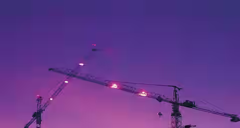 A construction site at dusk with illuminated cranes and structures. The sky is a gradient of pink and purple hues. Several cranes are actively working on a large cylindrical building in progress, surrounded by scaffolding and other partially built structures.