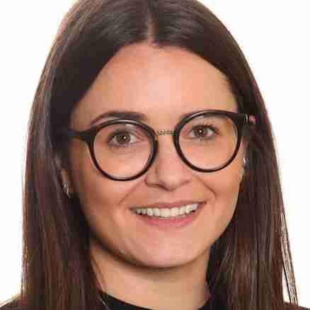 A woman with long straight brown hair, wearing black-rimmed glasses and a black top. She is smiling and wearing a necklace with a name pendant. The background is plain white.